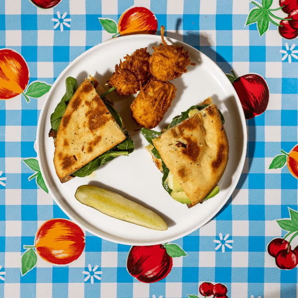 A white plate with two quesadilla halves, fried fritters, and a pickle spear on a blue checkered tablecloth.