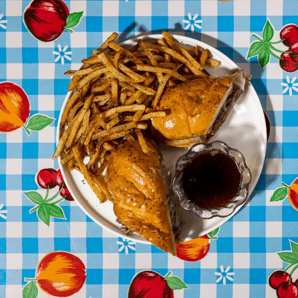 A Doo-Dah Diner French Dip sandwich with fries and au jus on a blue and white checkered tablecloth.